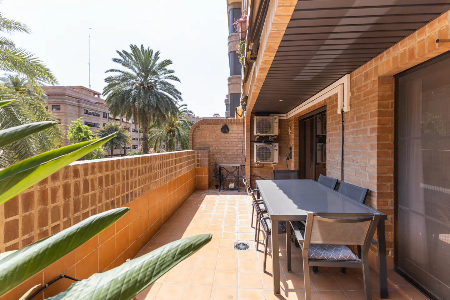 A sunny patio with a gray table and chairs on a tiled floor, brick walls, and palm trees in the background.