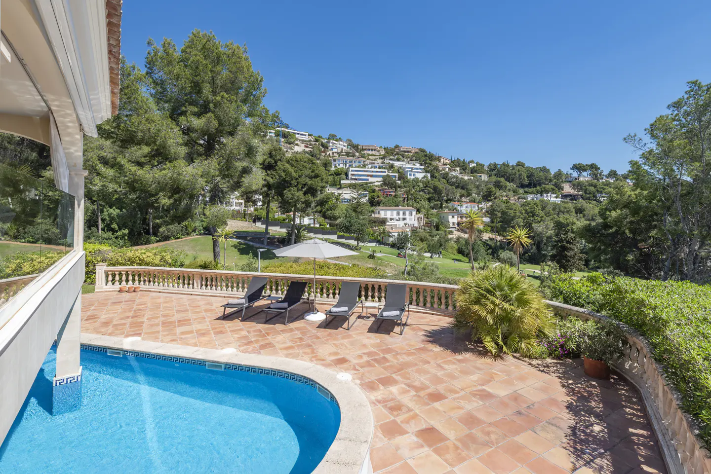 Outdoor patio with a blue pool, terracotta tiles, and four lounge chairs overlooking a green hillside with houses.