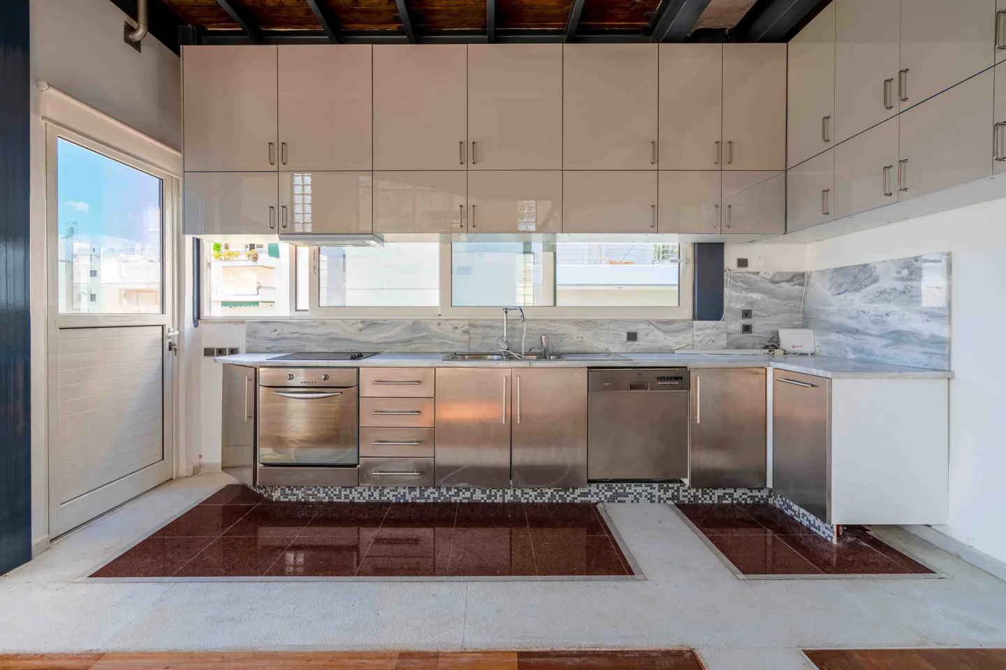 A modern kitchen with stainless steel appliances, white cabinets, and brown tile flooring. A window overlooks a cityscape.