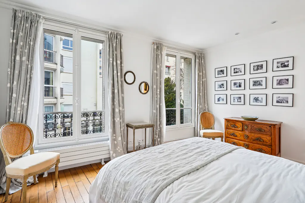 Bright bedroom with hardwood floors, white walls, and two windows with floral curtains. A bed, dresser, and framed photos complete the scene.