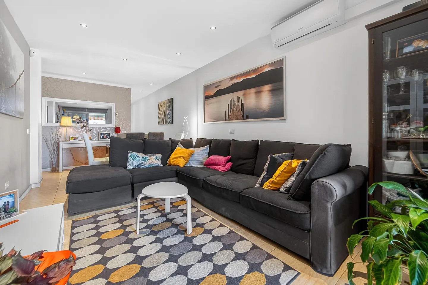 Living room with a large gray sectional sofa, colorful pillows, and a patterned rug. A white table and artwork decorate the space.