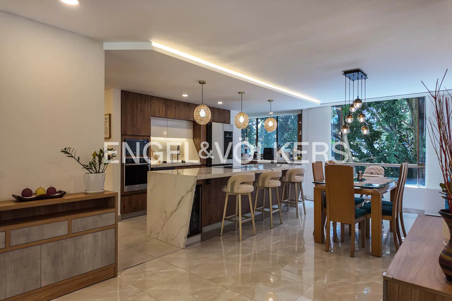 Open-concept kitchen and dining area with marble island, wood cabinets, and large windows overlooking greenery.