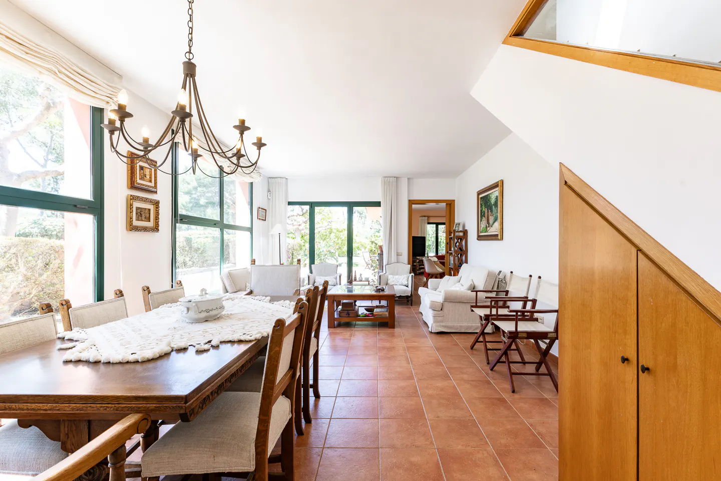 Bright, open-plan living and dining area with terracotta tile flooring. A large wooden dining table sits under a chandelier, with a living room beyond.