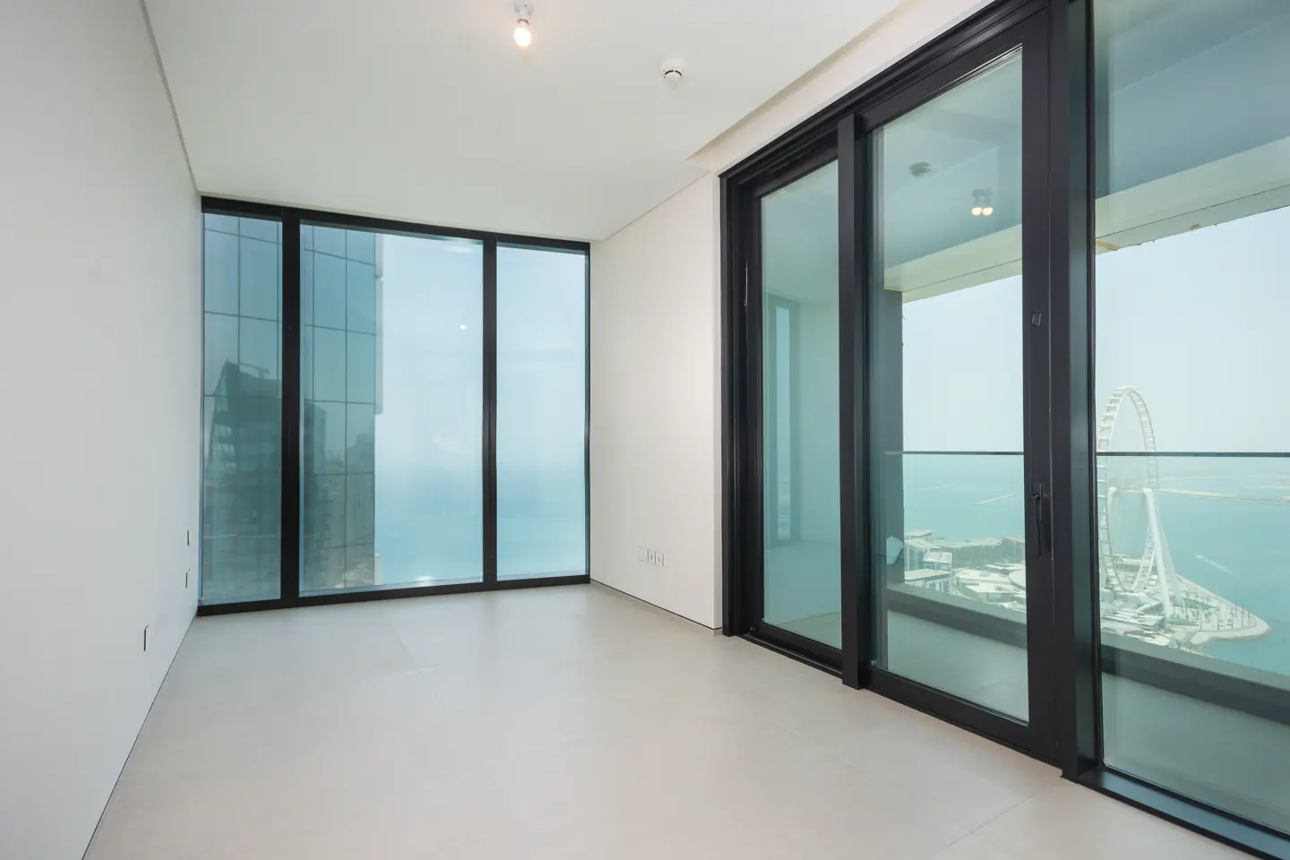 Empty room with white walls, light floor, and floor-to-ceiling windows. Ocean view with a Ferris wheel visible.