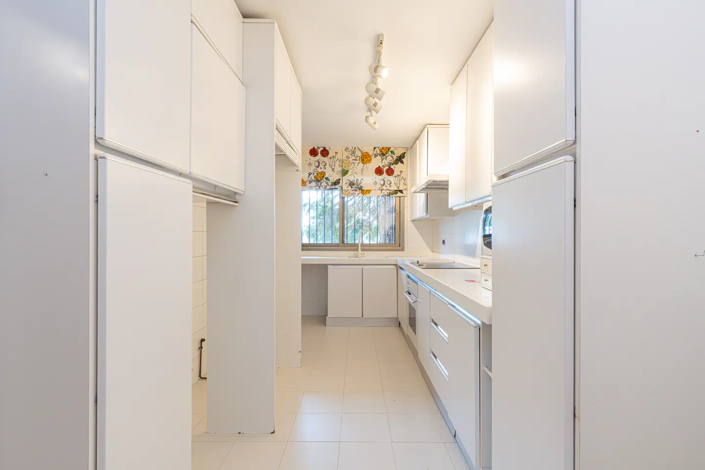 A bright, narrow kitchen with white cabinets, counters, and tile floors. A window with a floral valance is at the end of the kitchen.