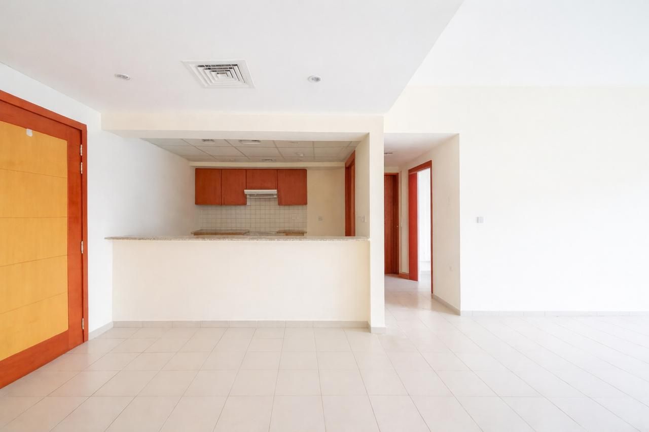 An empty room with tile floors, white walls, and a kitchen area with brown cabinets. A doorway with a red frame is visible.