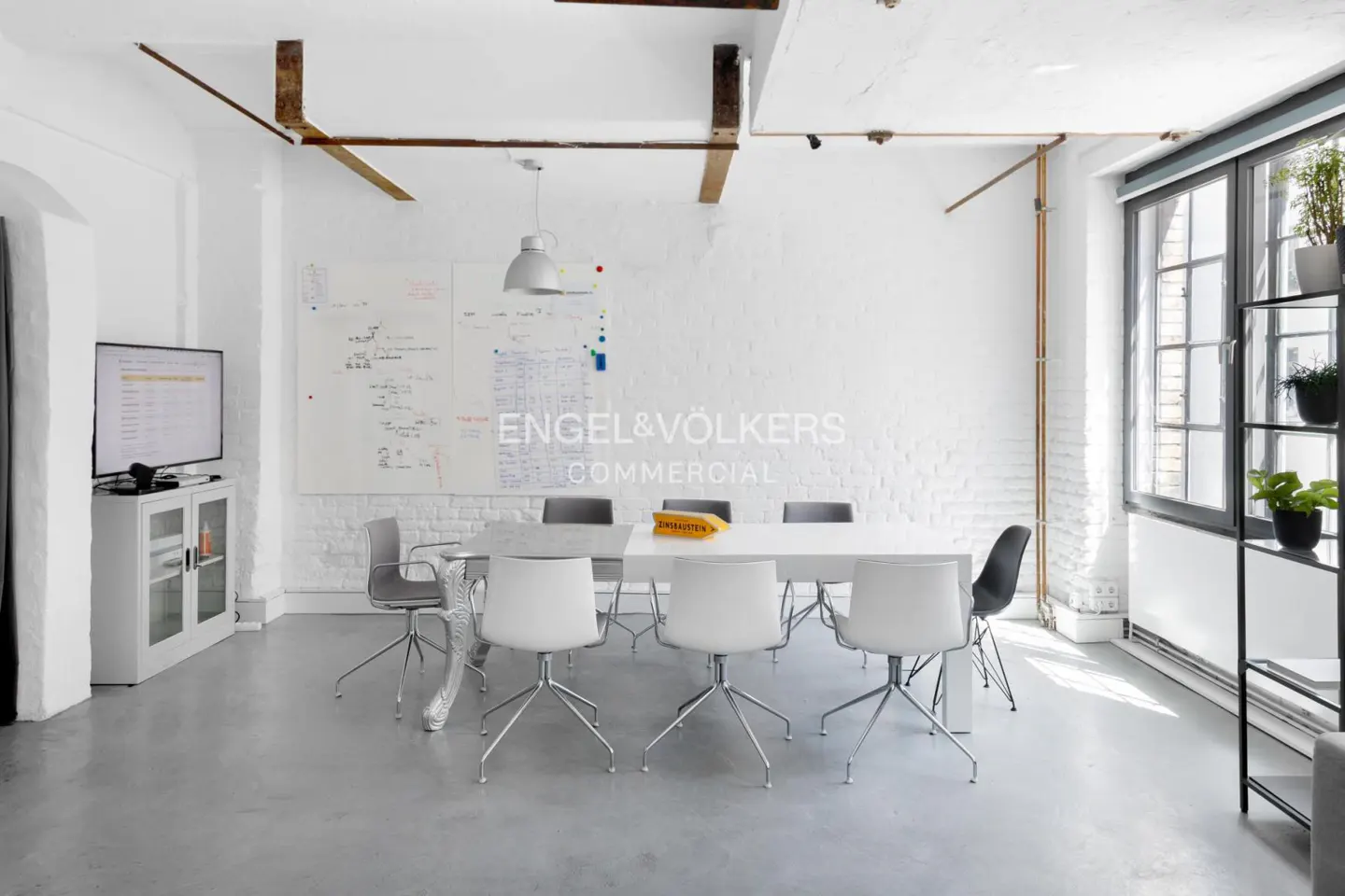 Bright, modern office space with a white table, chairs, and whiteboard. Engel & Völkers logo on the wall. Shelves with plants near the window.