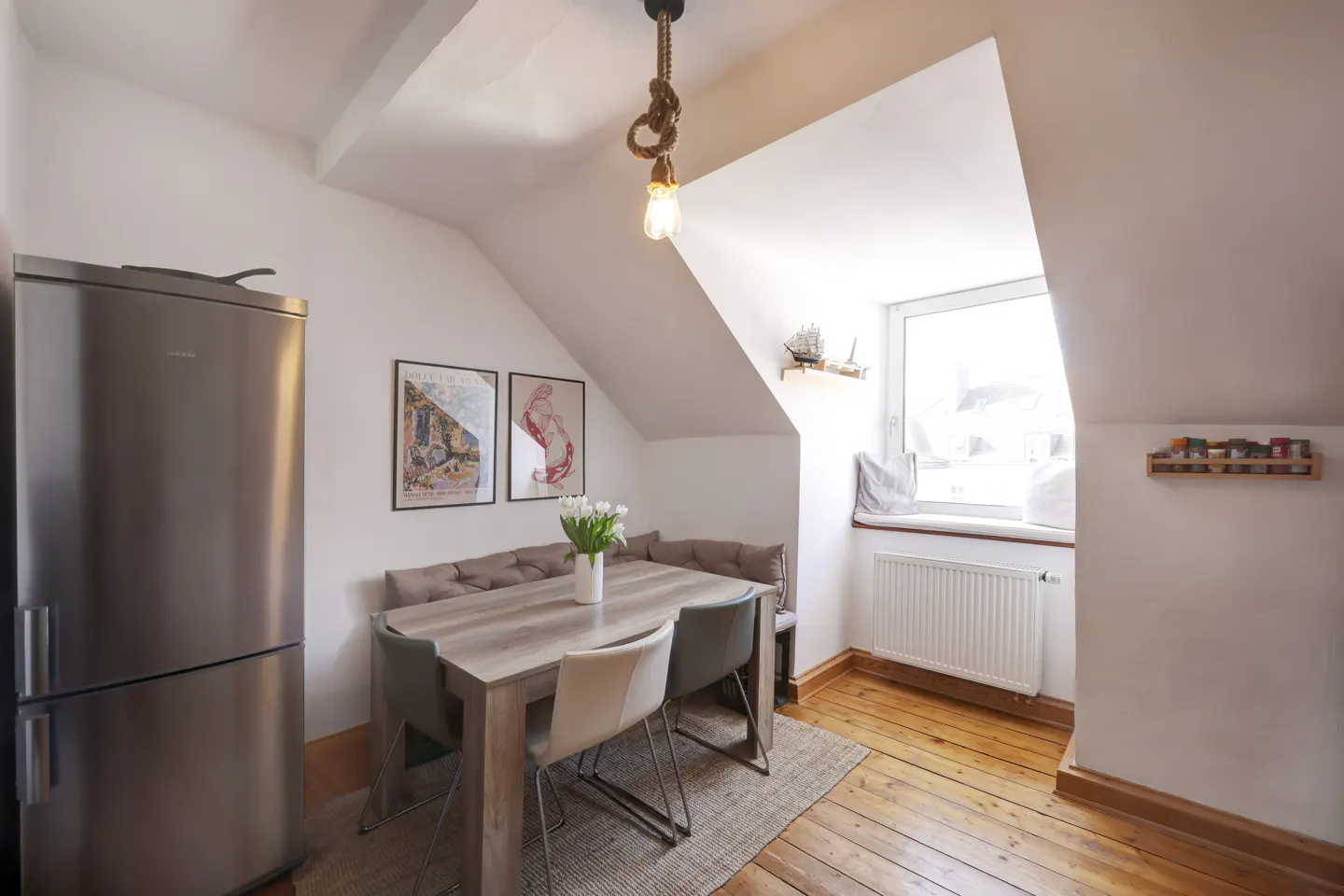 Attic dining area with a stainless steel fridge, wood table, bench seating, and white walls. A rope light fixture hangs above.