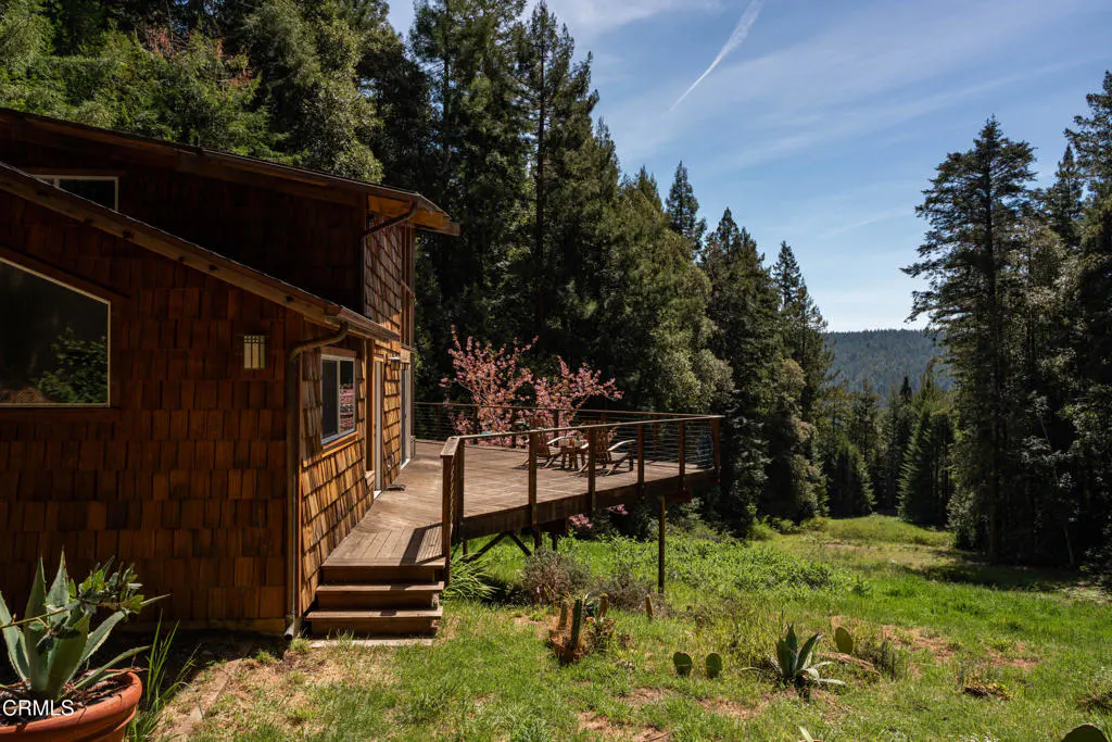 Exterior view of a wood-shingled house with a deck overlooking a green meadow and forest. Blue sky with wispy clouds above.