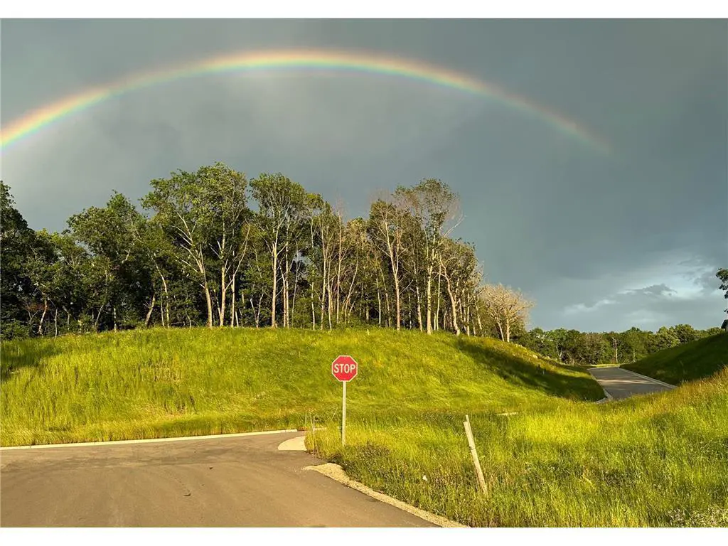A rainbow arches over a grassy hill with trees, a road, and a stop sign.