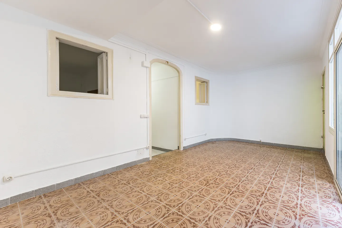Bright, empty room with white walls, brown patterned tile floor, and two windows. An arched doorway leads to another room.