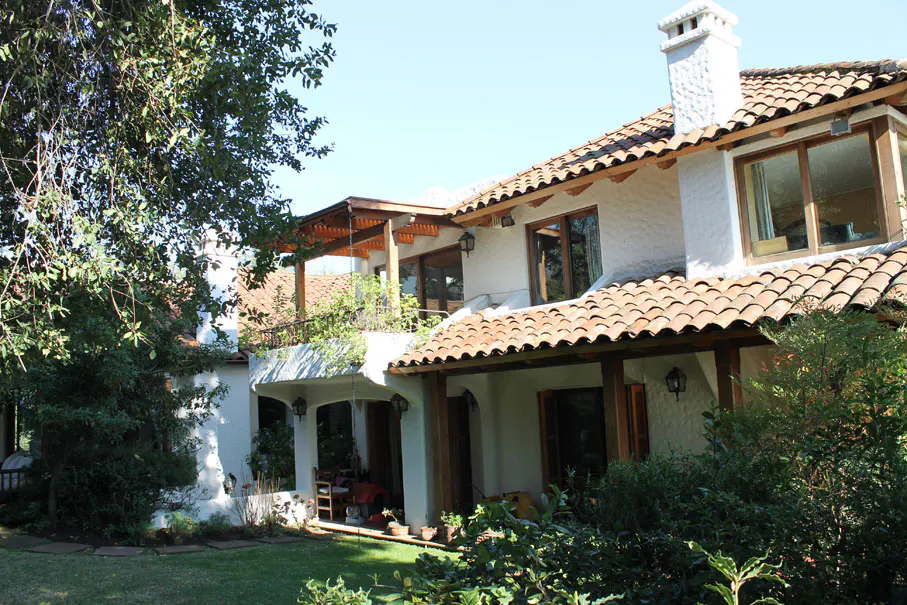 Exterior view of a two-story white house with a red tile roof, wooden beams, and a chimney, surrounded by green trees and bushes.
