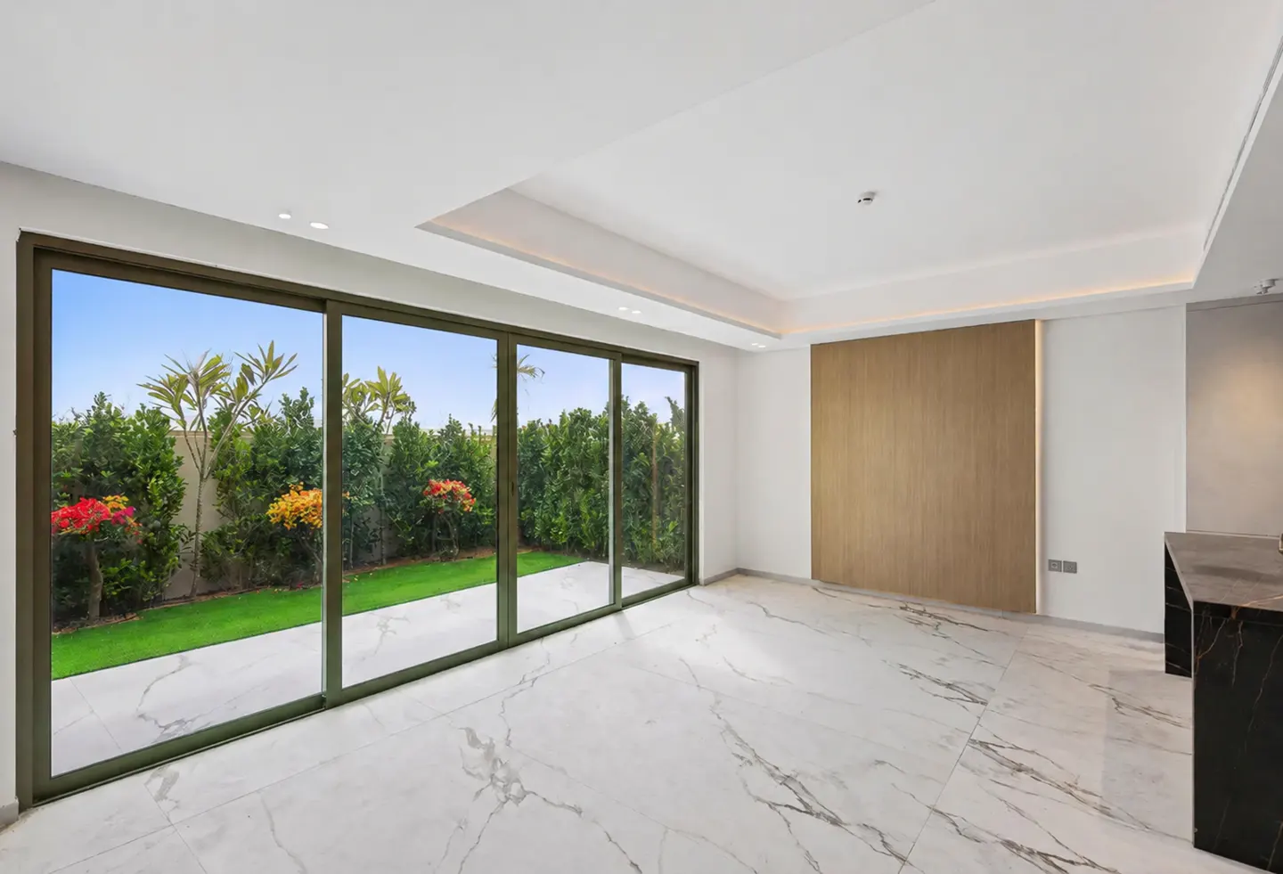 Bright, empty room with marble floors and a large sliding glass door to a green garden. A wood panel accents the wall.