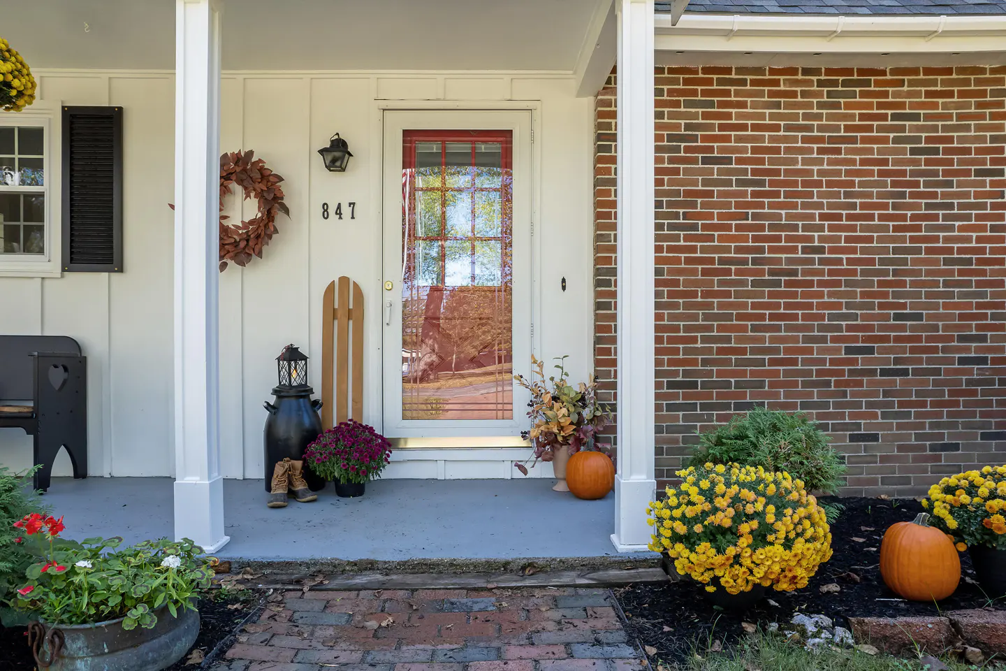 Front porch of a white house with red brick. Fall decorations include mums, pumpkins, and a wreath.