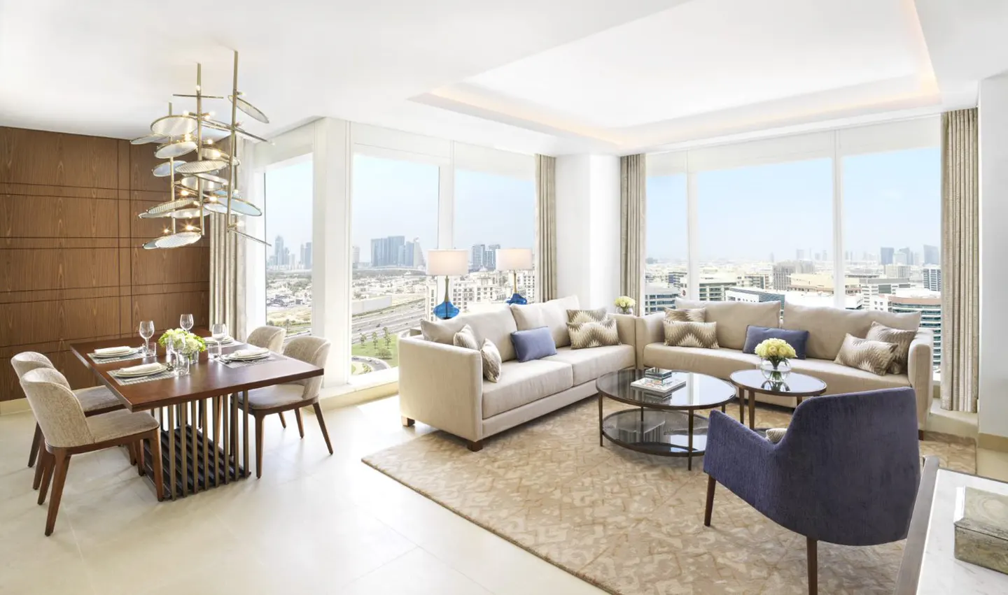 Modern open-plan living and dining area with floor-to-ceiling windows, beige sofas, a dark wood table, and city views.