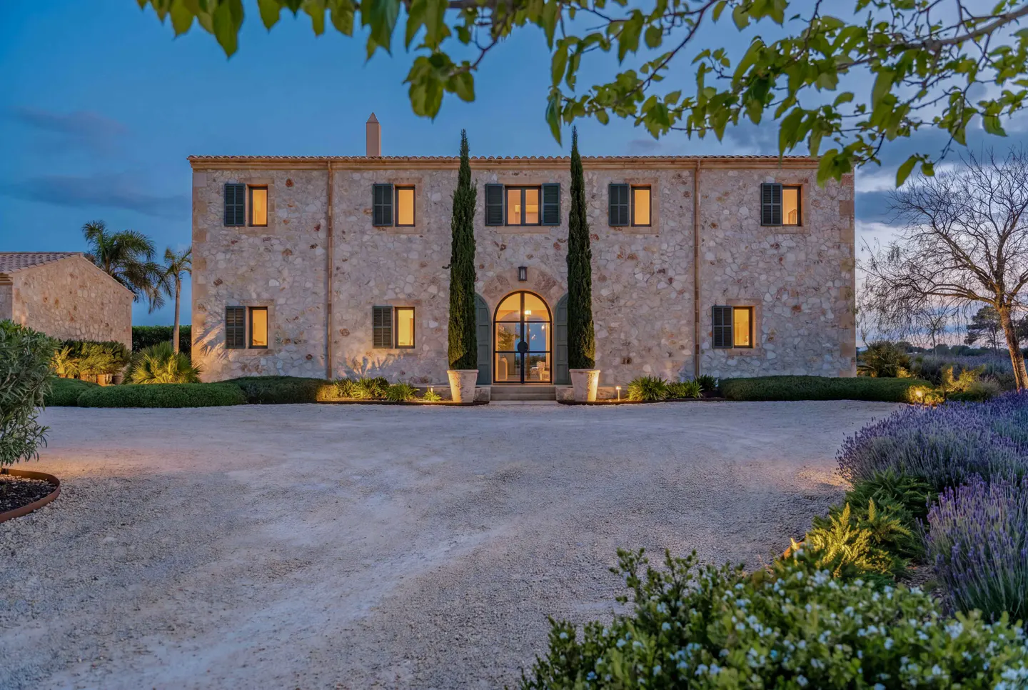 Two-story stone house with dark green shutters and arched doorway at dusk. Tall cypress trees flank the entrance. Gravel driveway and manicured landscaping.
