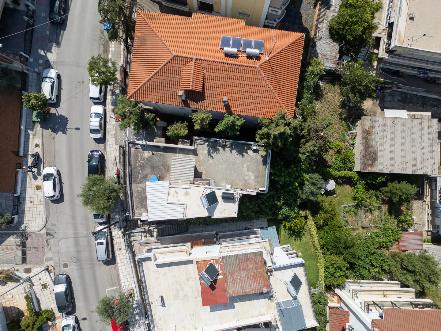 Aerial view of a street with parked cars next to houses featuring orange tiled roofs and solar water heaters.