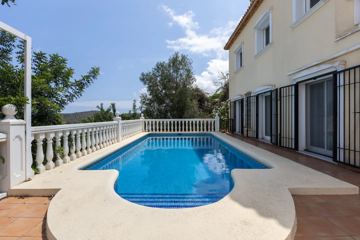 A rectangular swimming pool with blue water sits on a tiled patio next to a cream-colored house with a white balustrade.