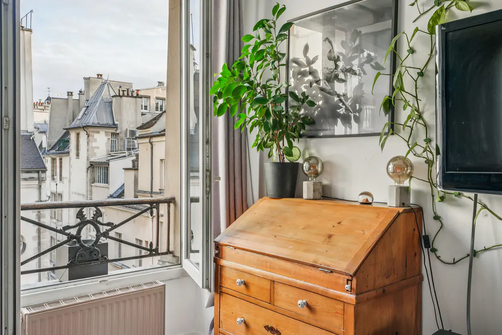 Bright room with open window showing Paris rooftops. A wooden desk with lamps and plants sits against a white wall with art and a TV.
