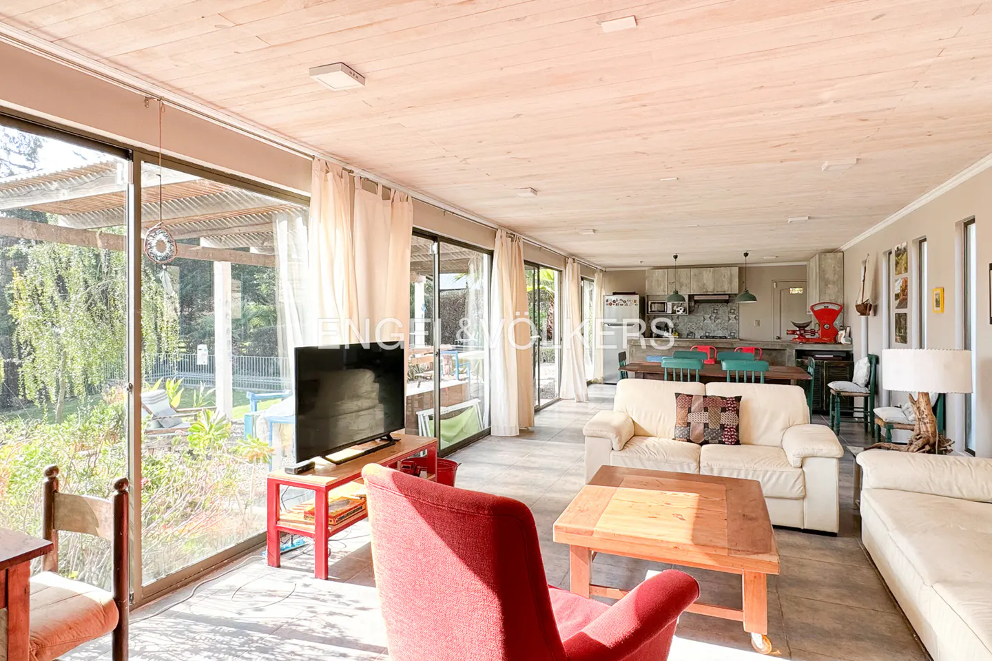 Bright living room with a red armchair, white sofa, wooden coffee table, and large glass doors overlooking a garden.