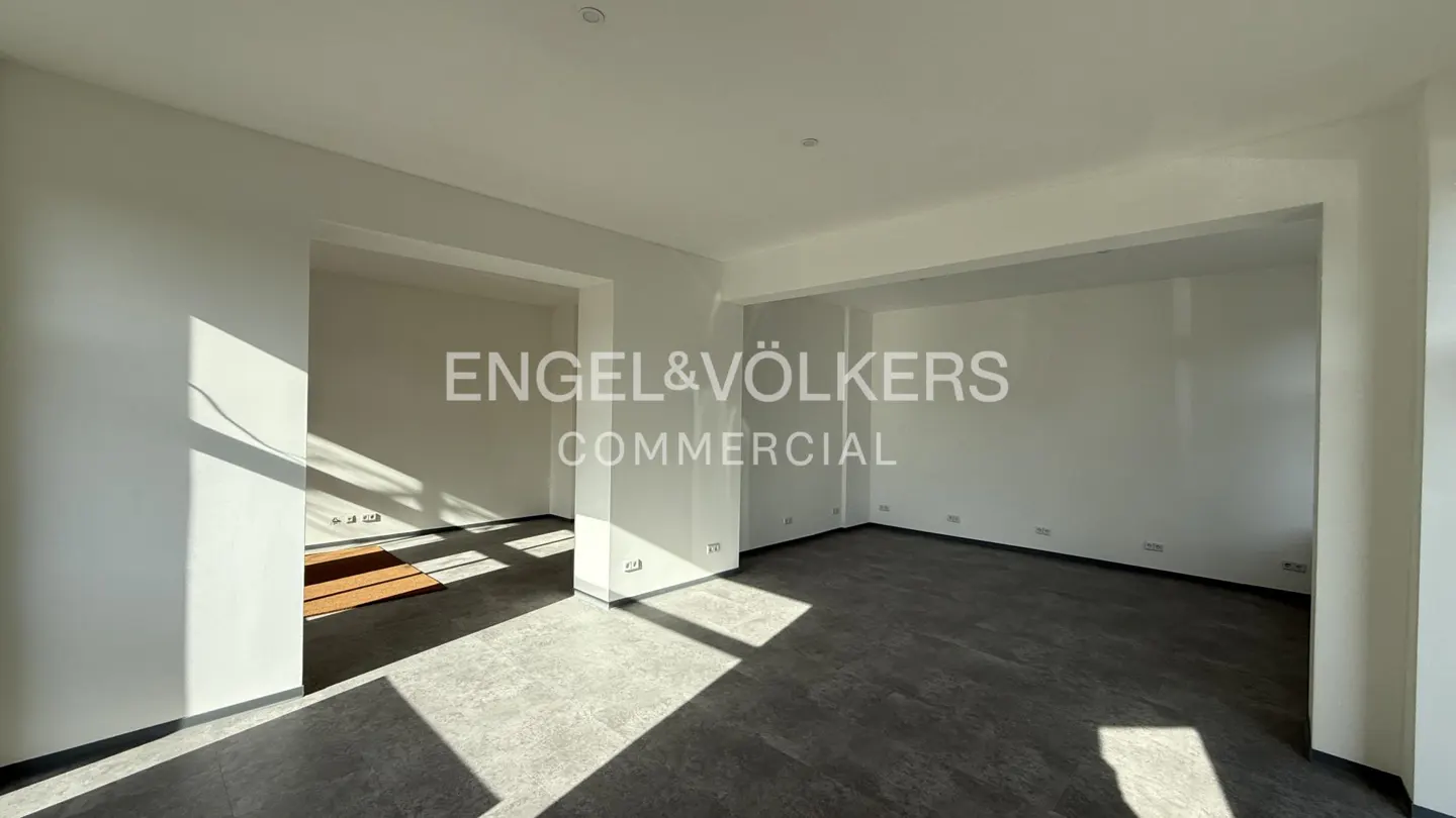 Empty commercial space with white walls, gray floor, and natural light. Engel & Völkers logo in the center.