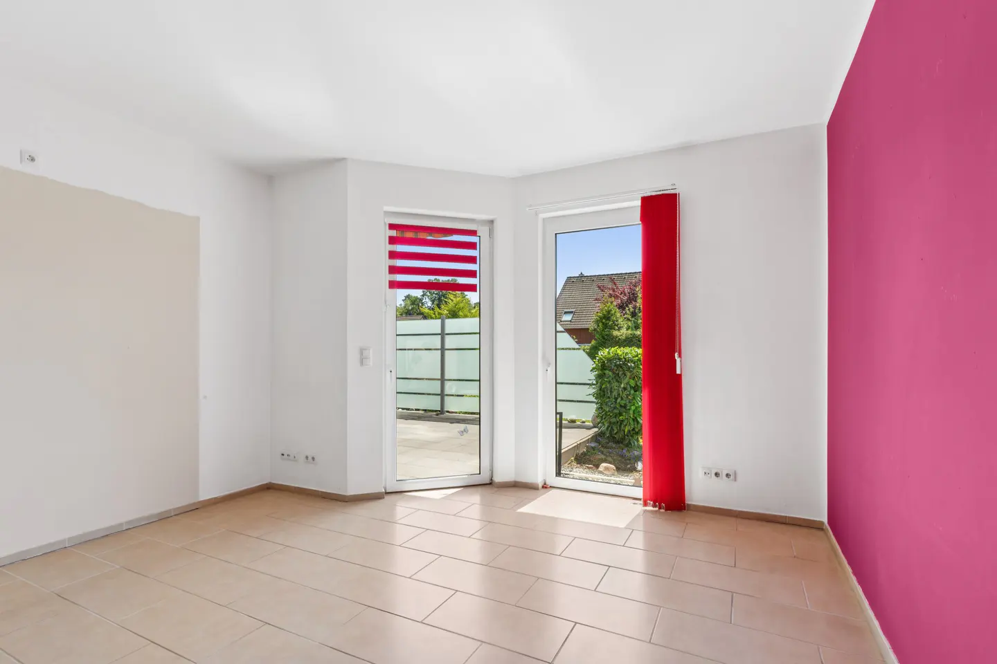 Bright, empty room with beige tile floor, white walls, and a pink accent wall. Two glass doors lead to a patio with greenery. Red blinds adorn the windows.
