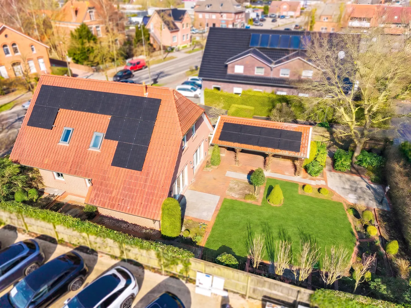 Aerial view of a house with solar panels on a red tile roof, a green lawn, and several cars parked in front.