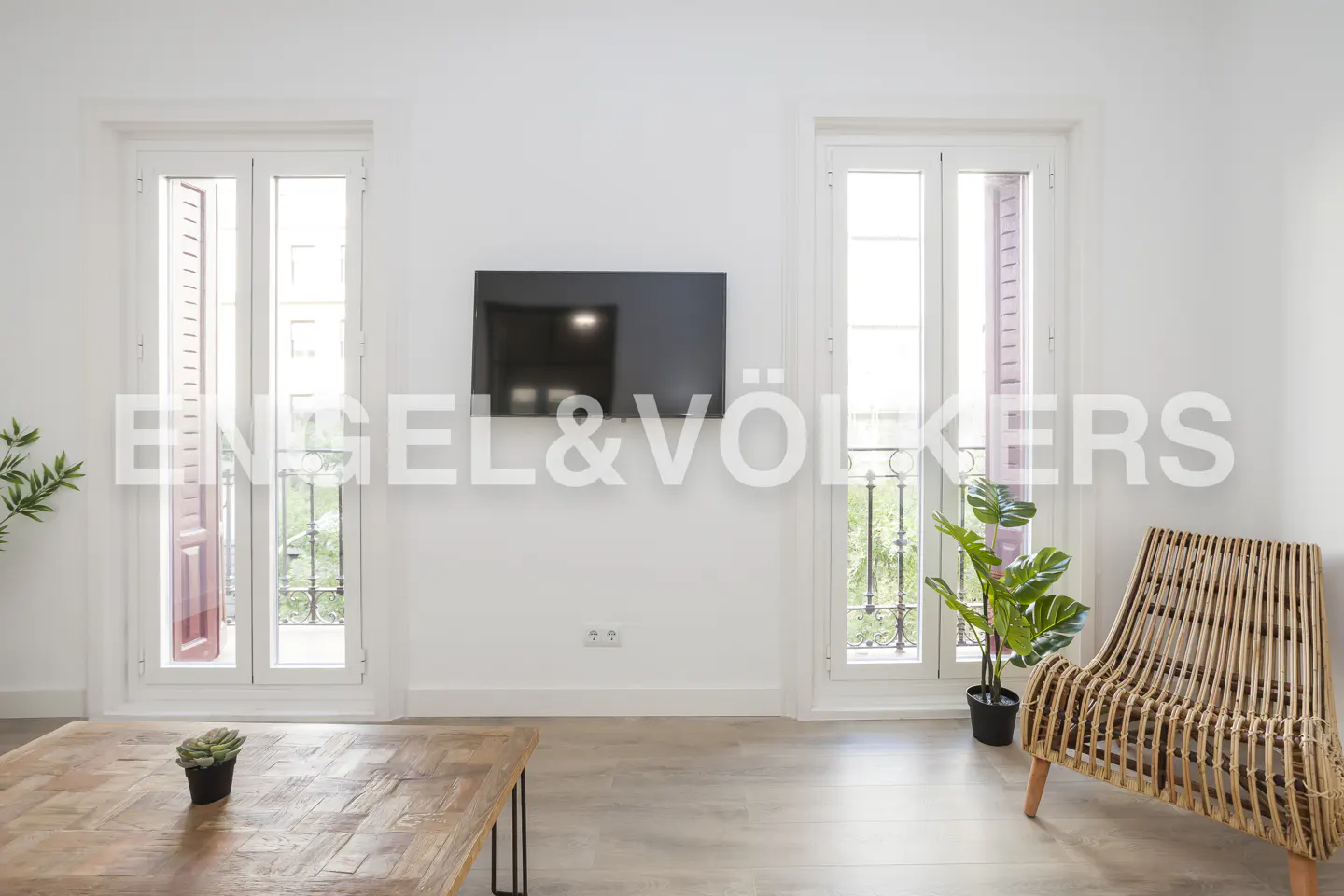 Bright living room with white walls, a wall-mounted TV, two balcony doors, a wooden coffee table, and a rattan chair.
