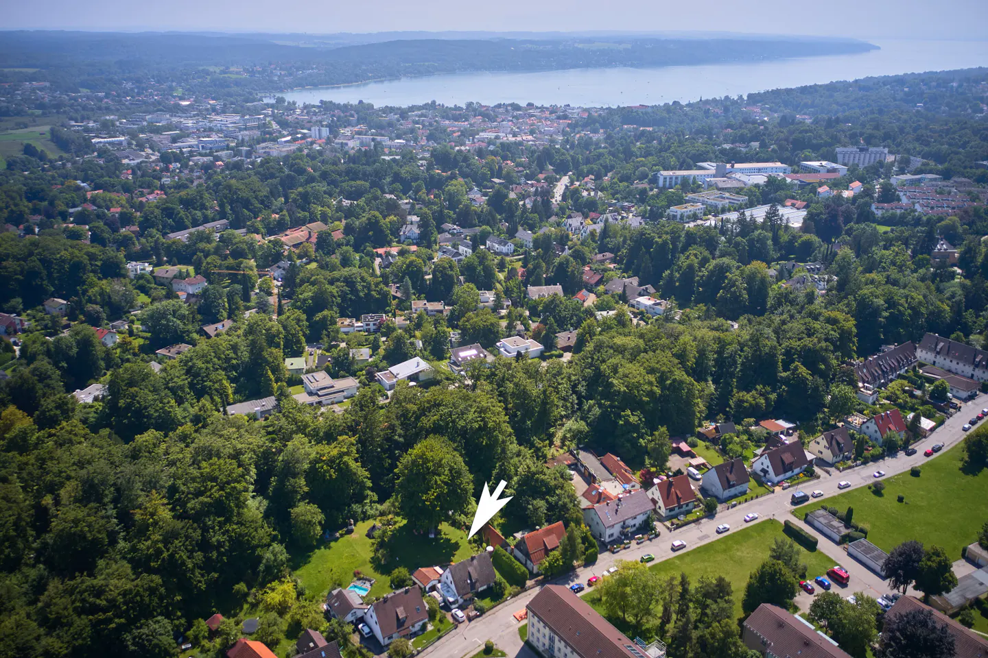 Aerial view of a neighborhood with green trees, houses with red roofs, and a lake in the background. An arrow points to a house.