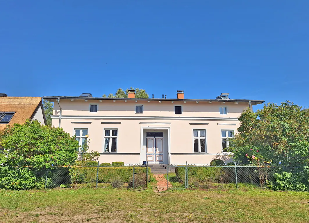 Two-story beige house with white trim, a brick walkway, and a green lawn under a clear blue sky.