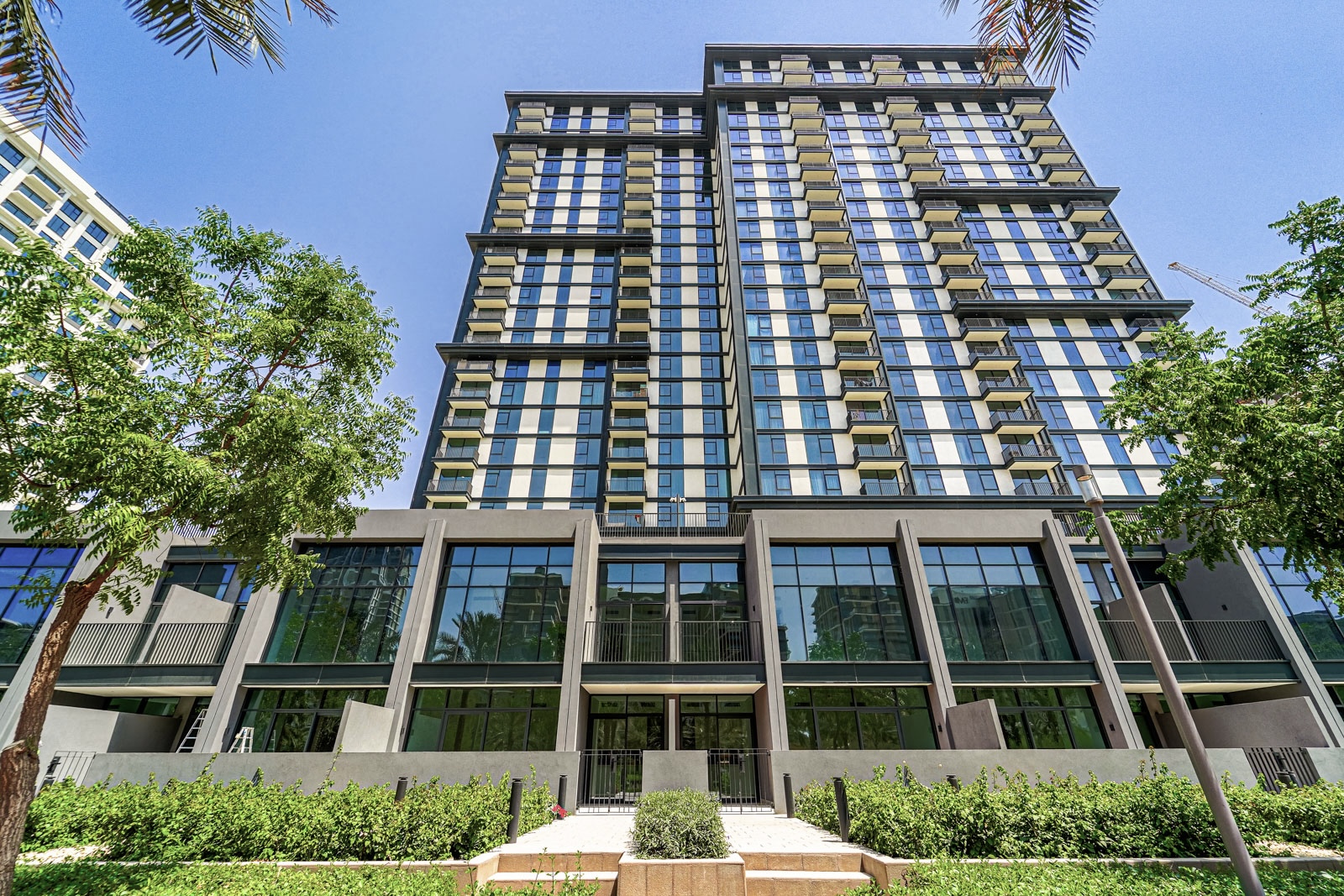 Low-angle view of a modern high-rise apartment building with glass balconies, dark frames, and lush green landscaping. [AI-generated text]