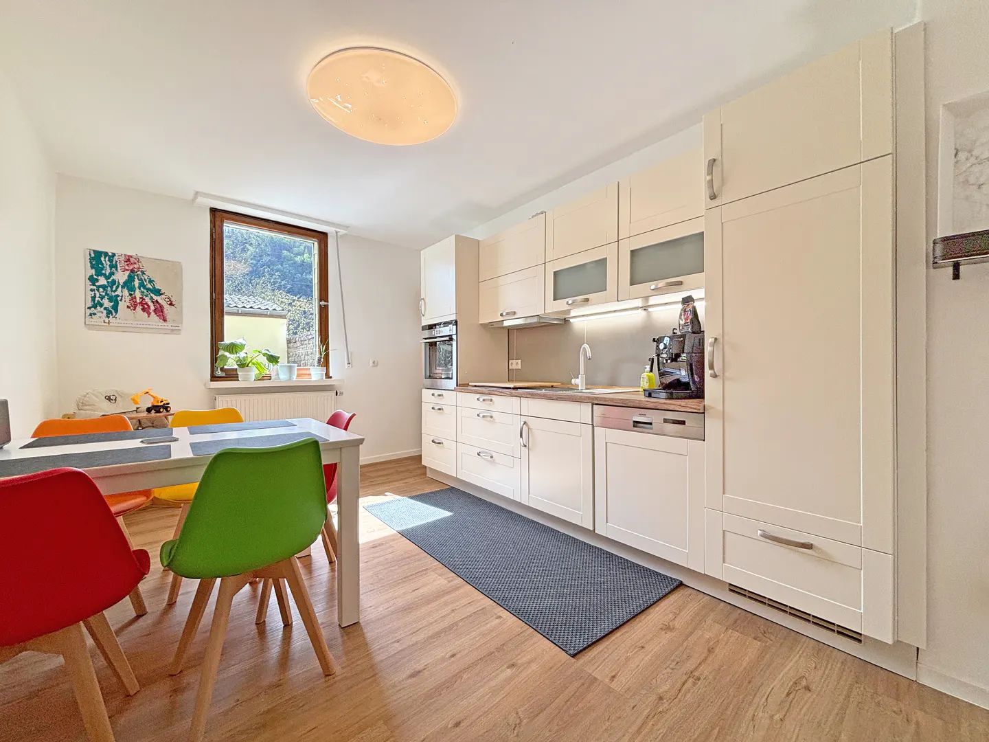 Bright kitchen with wood floors, white cabinets, and a dining table with colorful chairs. A window overlooks greenery.