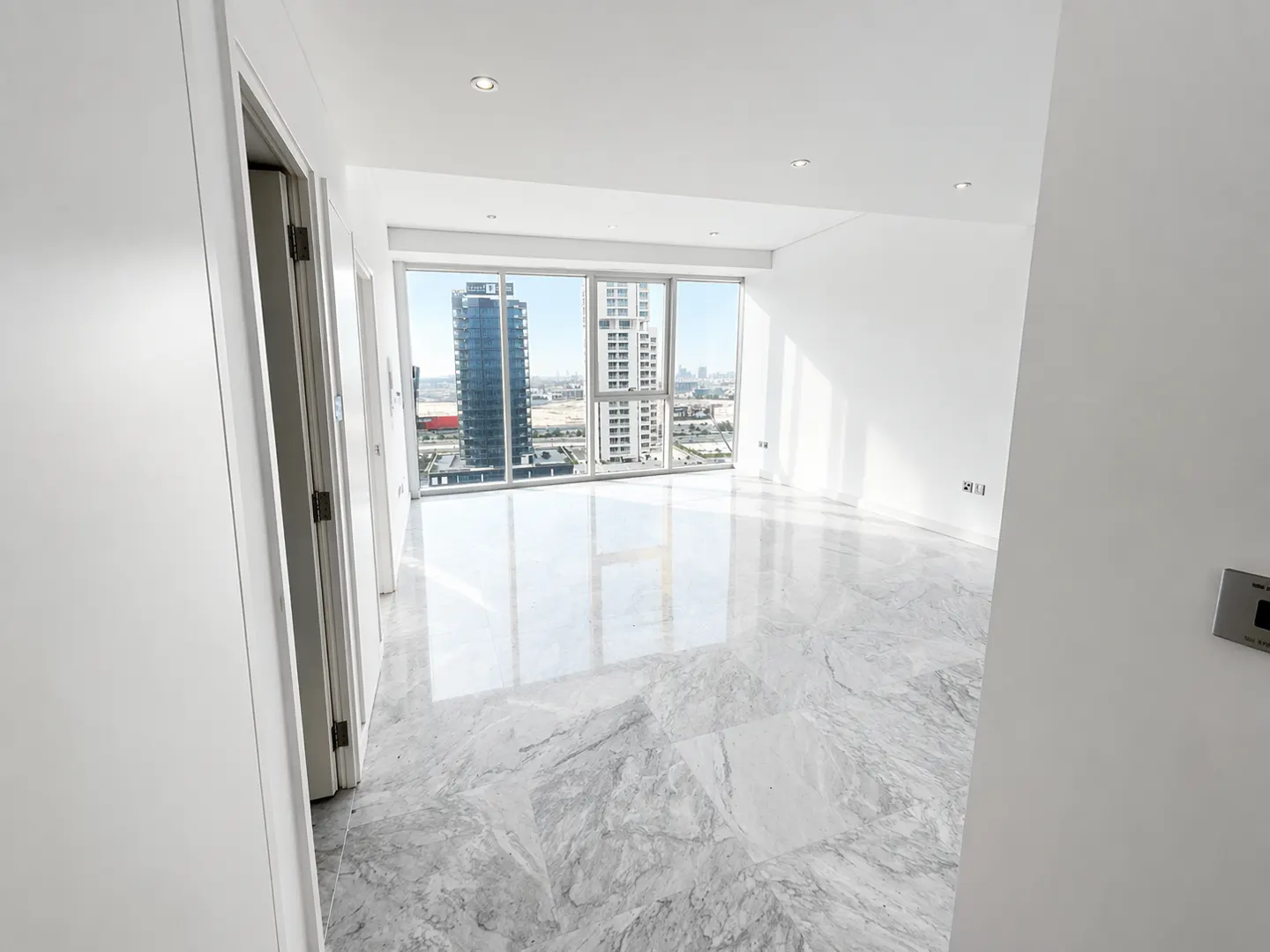 Bright, empty room with white walls and gray marble floors. Large windows offer a city view with tall buildings.