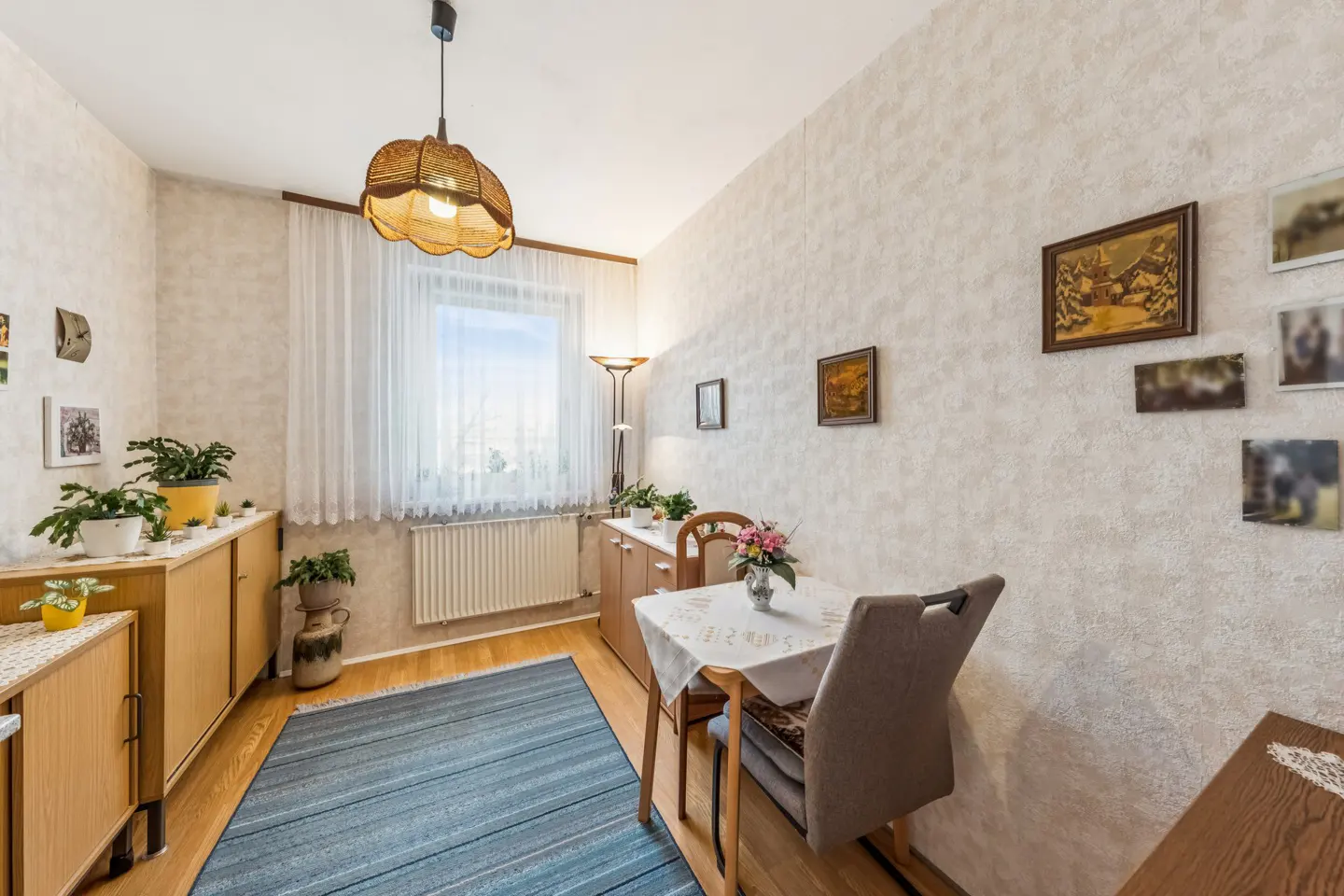 A cozy dining area with a table, chairs, and a blue rug. Light wood cabinets line the wall, adorned with plants. Pictures hang on the textured wallpaper.