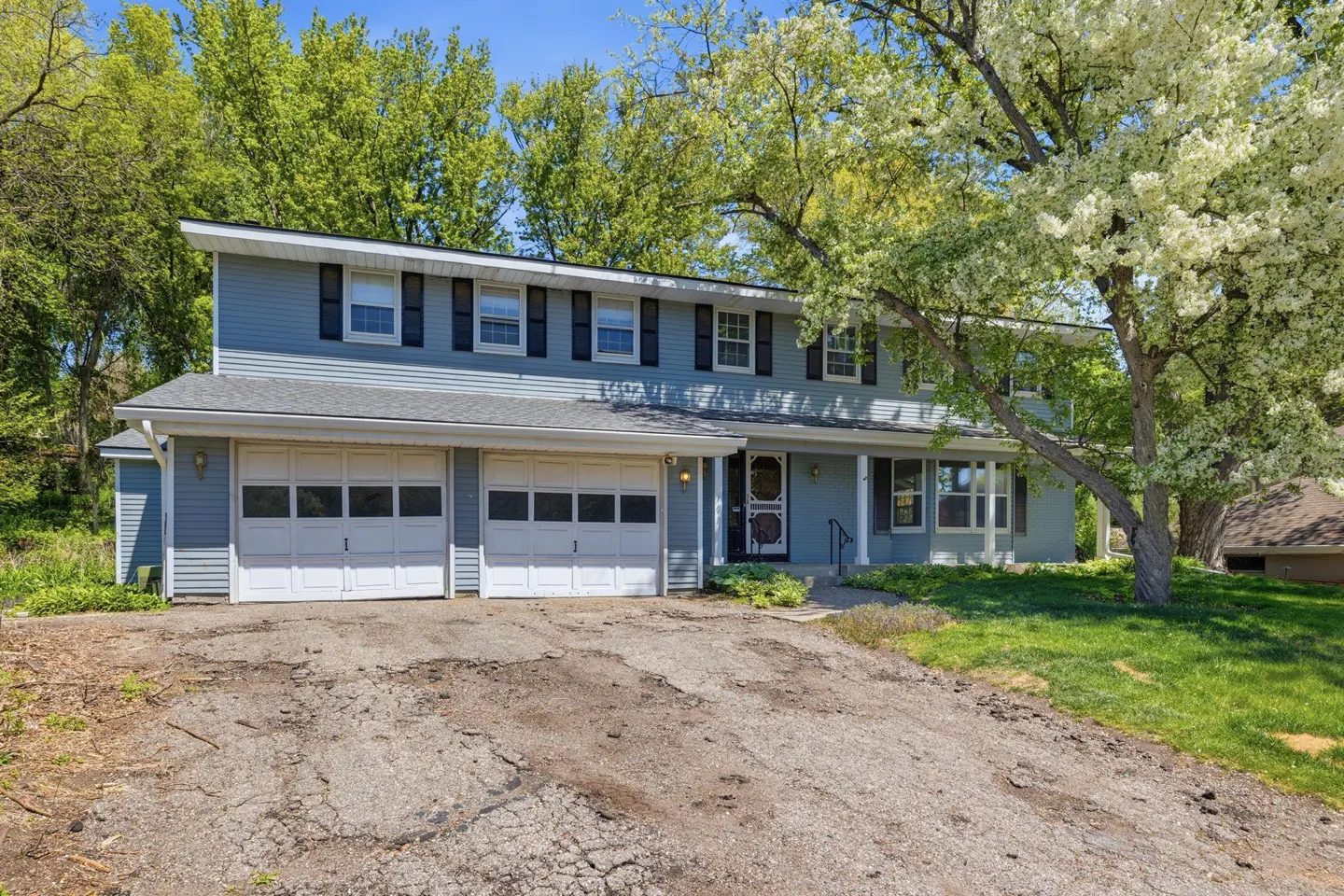 Two-story blue house with white garage doors, black shutters, and a cracked asphalt driveway. A flowering tree is on the right.