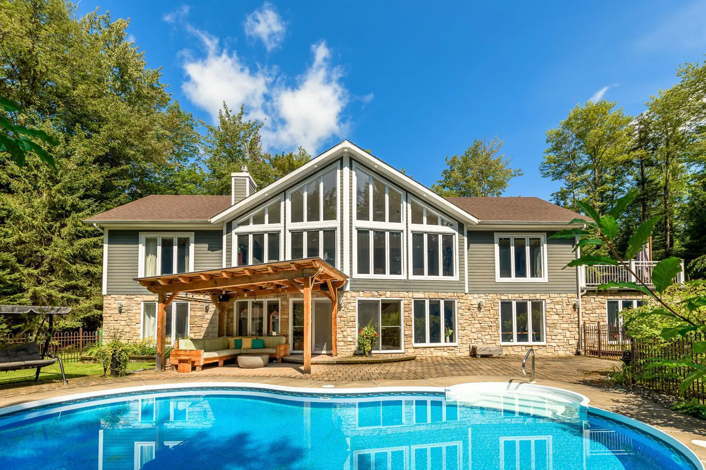 Two-story house with a pool. The house has gray siding, stone accents, and a wall of windows. A pergola covers an outdoor seating area.
