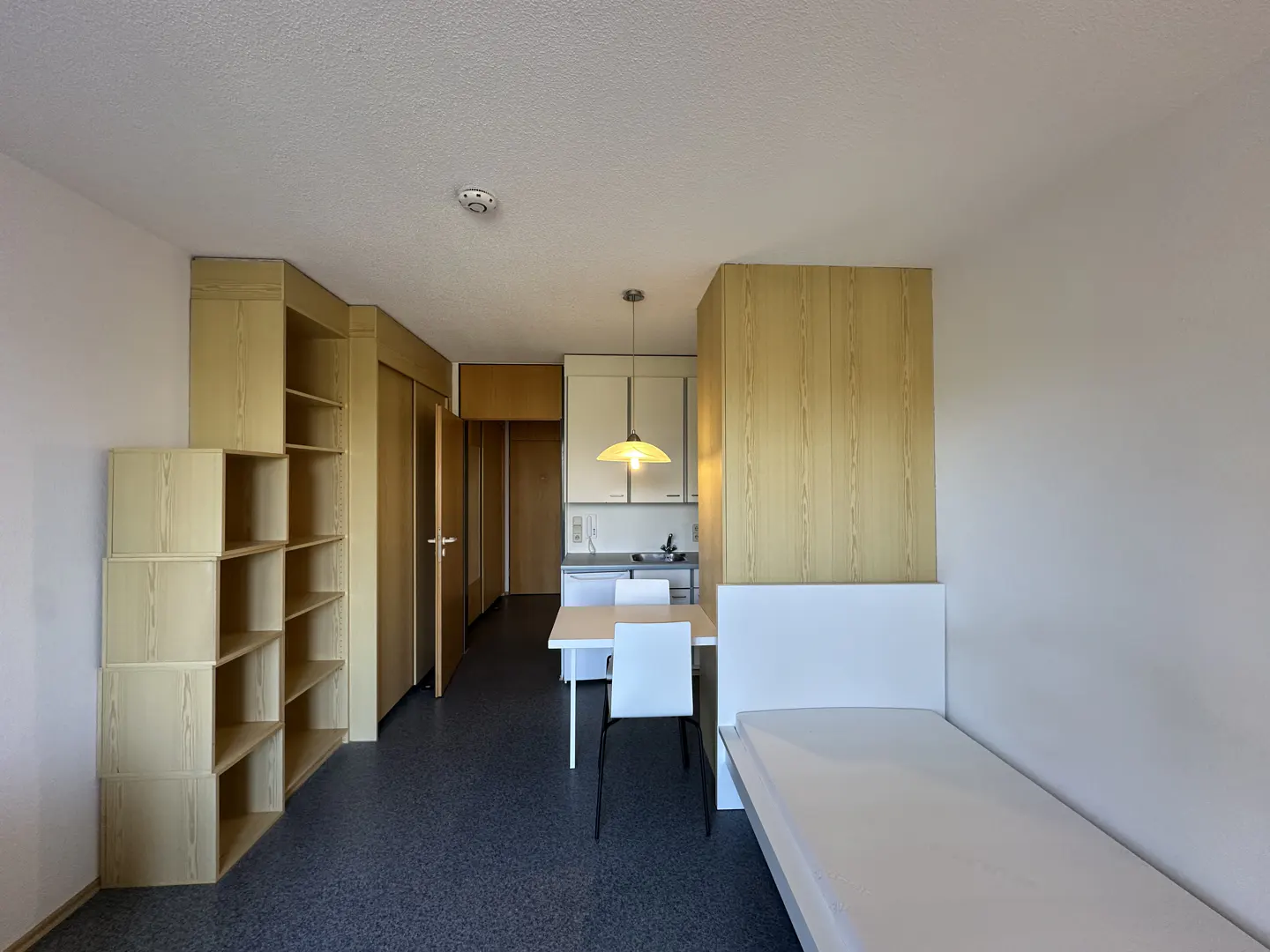 Studio apartment with a bed, table, chairs, and kitchenette. The walls are white, and the floor is dark gray. There is a wooden bookshelf.