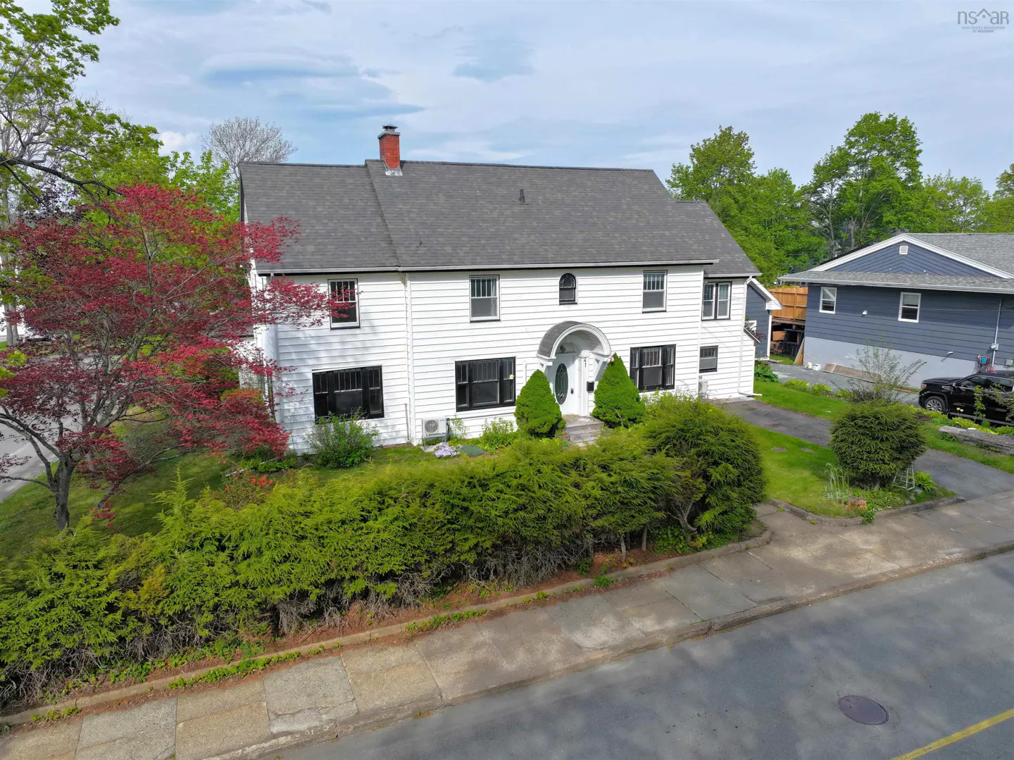 Two-story white house with black shutters, gray roof, and red chimney. Green bushes line the front. A red-leaved tree is on the left.