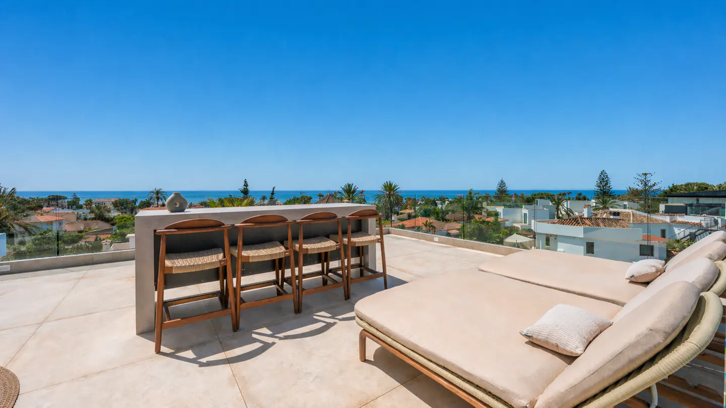 Rooftop patio with bar, stools, and lounge chairs overlooking the ocean on a sunny day.