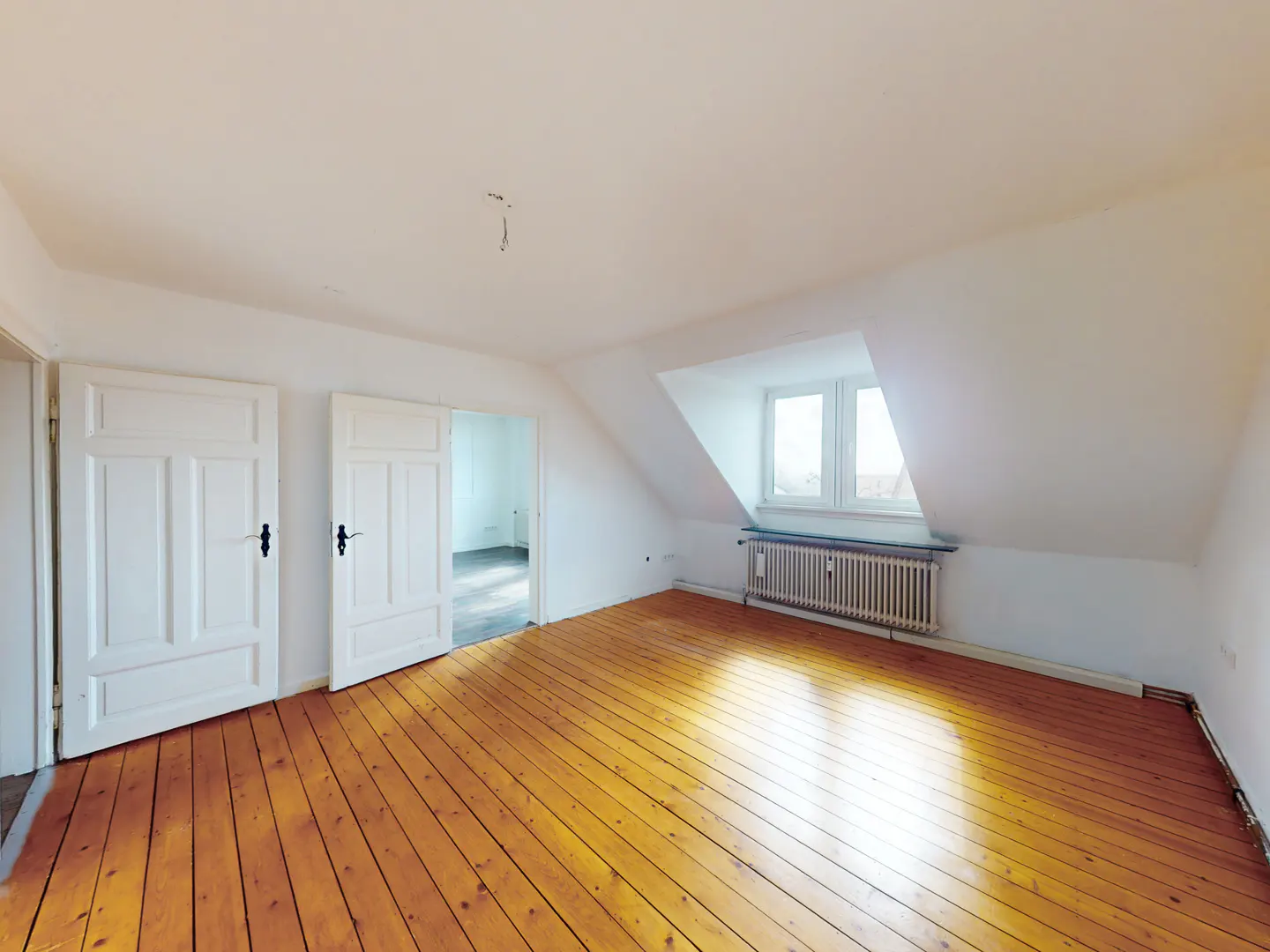 Attic room with white walls, wood floors, and a radiator under a window. Two white doors are on the left.