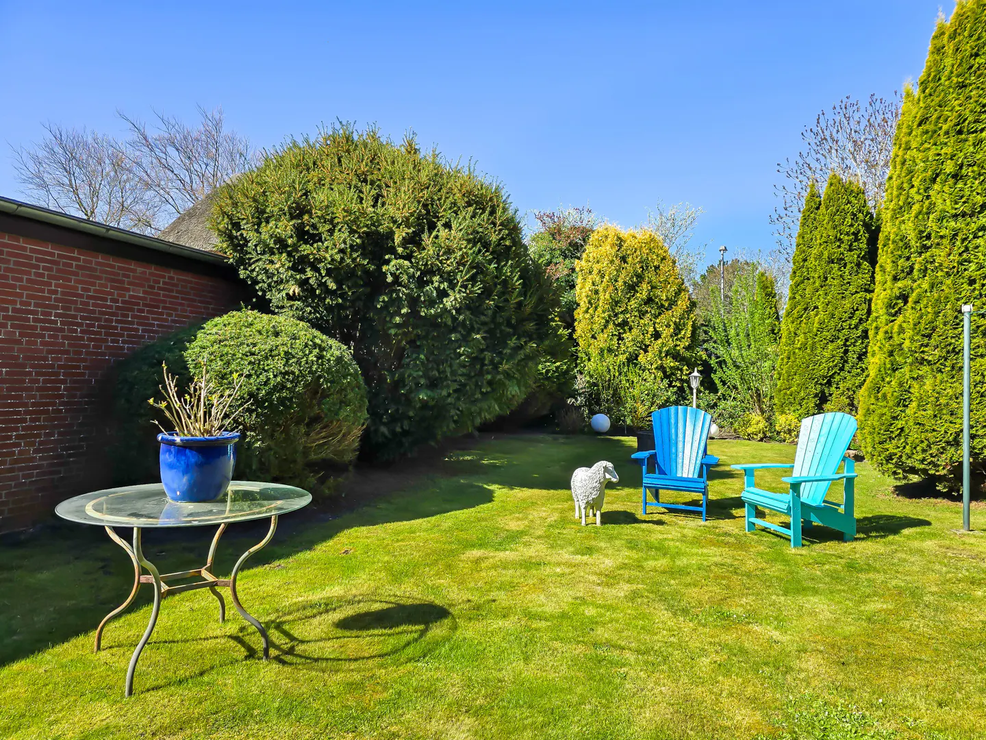 A lush green lawn with two blue Adirondack chairs, a sheep statue, and a glass table with a blue pot under a clear blue sky.