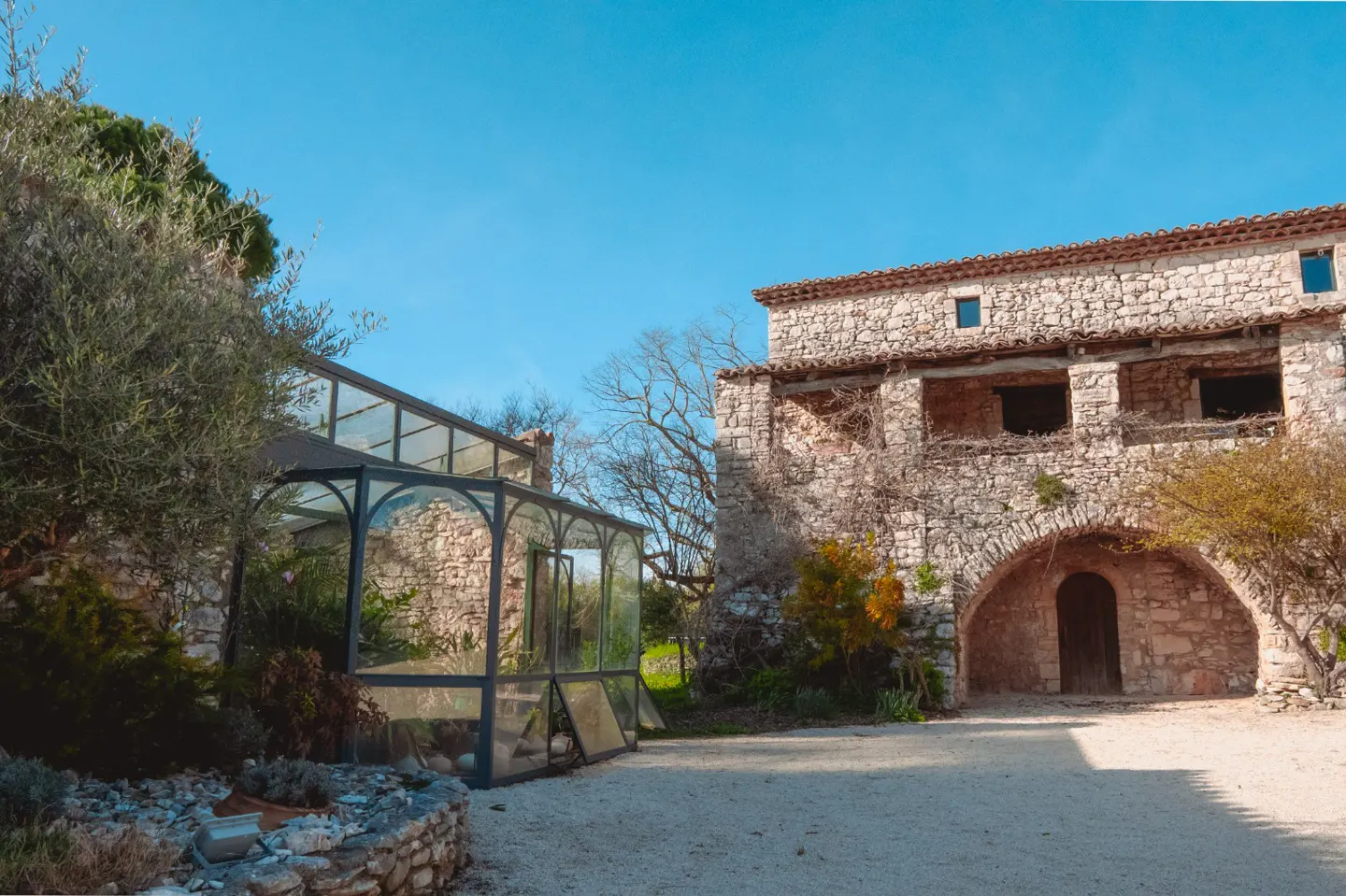 Exterior view of a stone building with arched entrance and a glass greenhouse on a gravel courtyard under a blue sky.