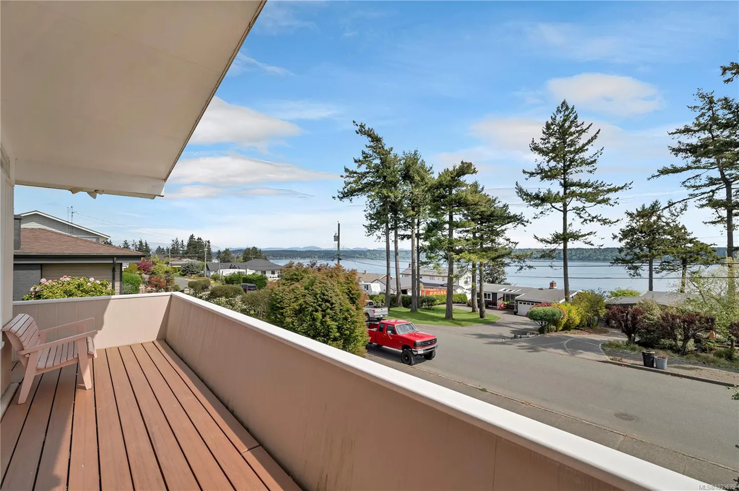 Balcony view with wooden deck and chair overlooking a street with a red truck, trees, houses, and a body of water under a blue sky.