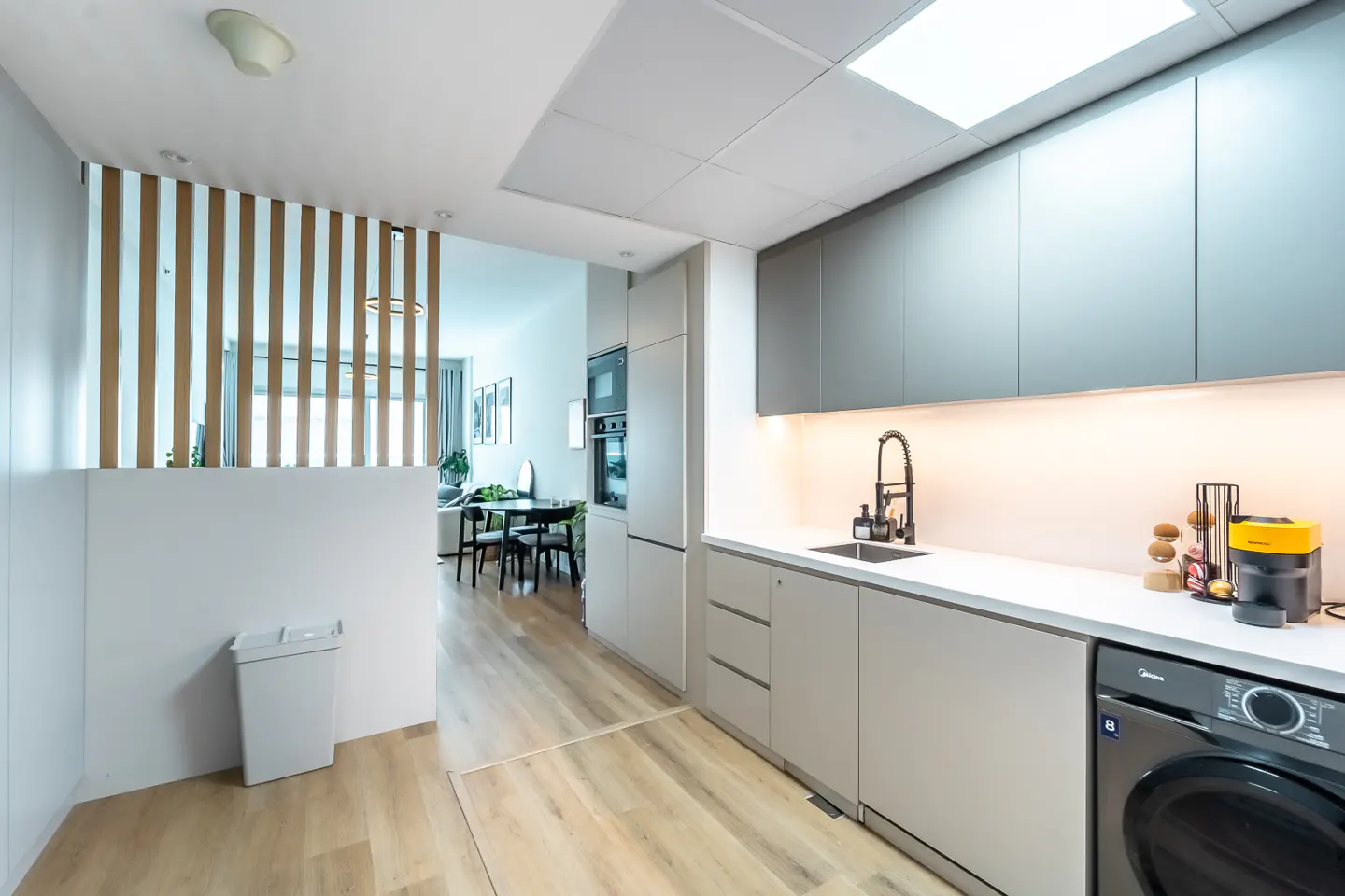 A modern kitchen with gray cabinets, white countertops, and a black faucet. A washing machine is visible on the right. A dining area is seen through a wooden slat partition.