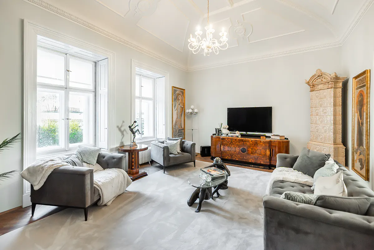 A bright living room with gray sofas, a unique coffee table, and a large TV on a wooden cabinet. Two large windows let in natural light.