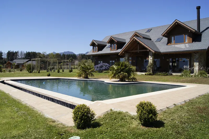 A large, rectangular pool sits in front of a wood and stone house with a gray roof on a sunny day.