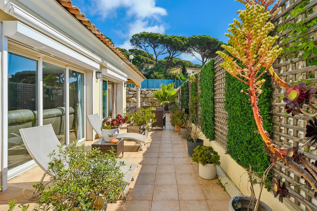 Outdoor patio with lounge chairs, table, and potted plants. Sliding glass doors lead to the interior. Green foliage and trees in the background.