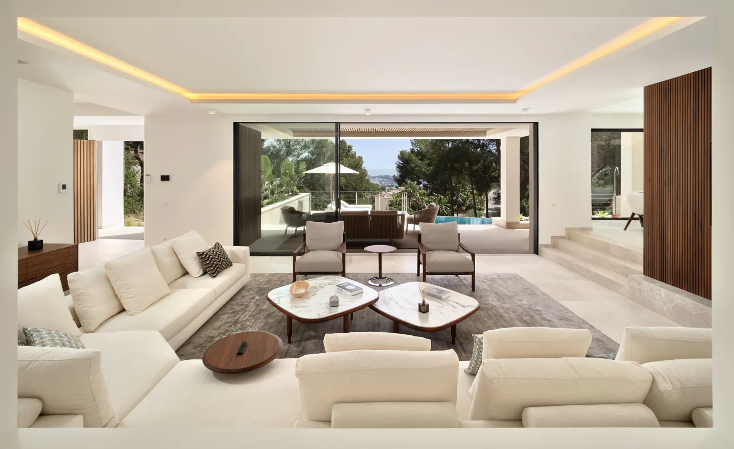Bright, modern living room with white sofas, chairs, and marble tables. Large sliding glass doors open to a patio with trees and a distant view.