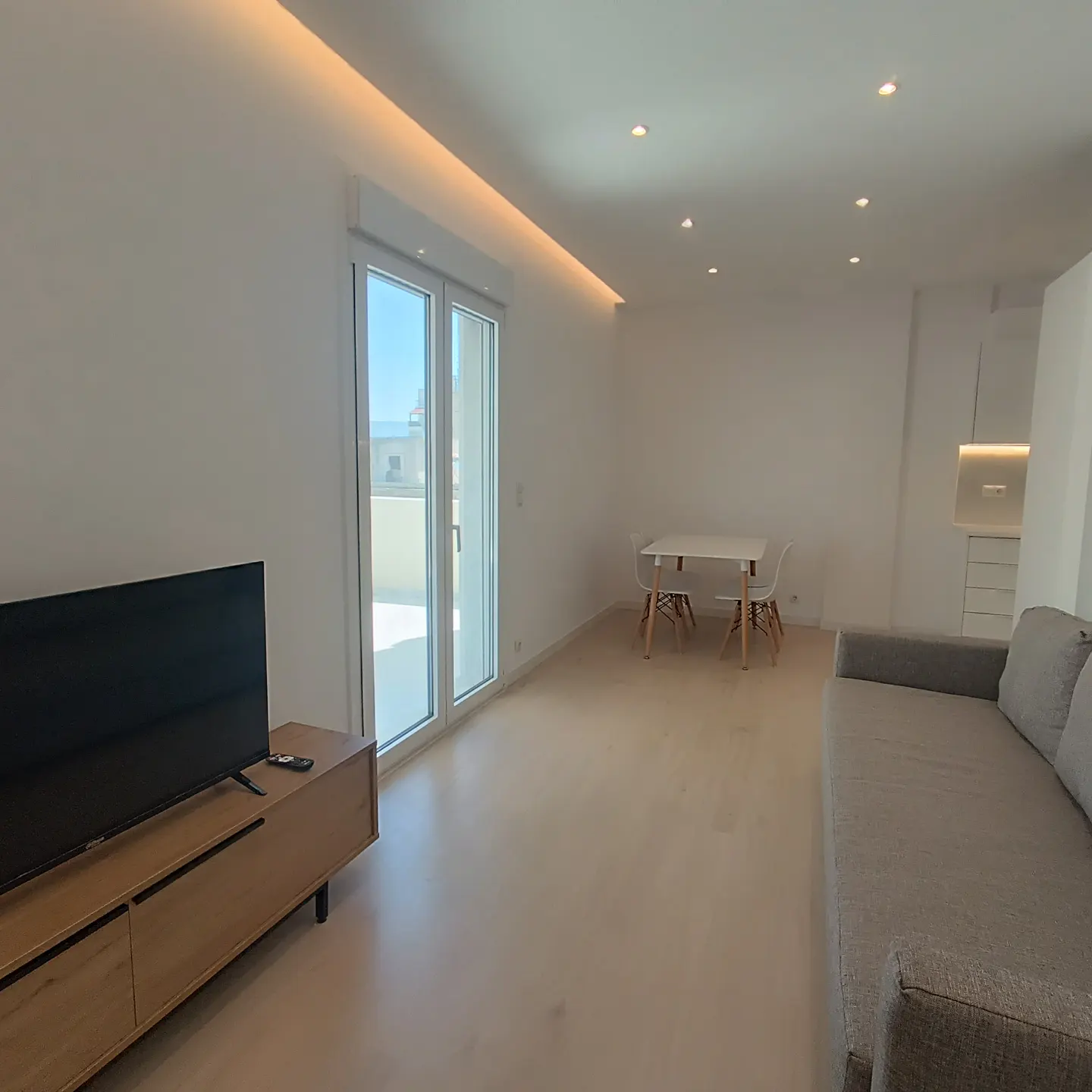 Bright, modern living room with white walls, light wood floors, and recessed lighting. A gray sofa, TV, dining table, and balcony doors are visible.