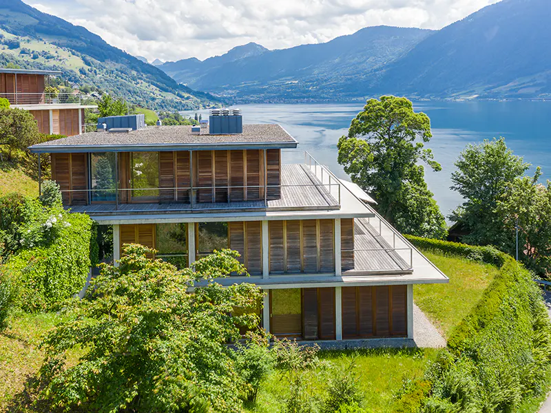 Modern multi-story wooden home with balconies overlooking a large lake and mountains on a sunny day.